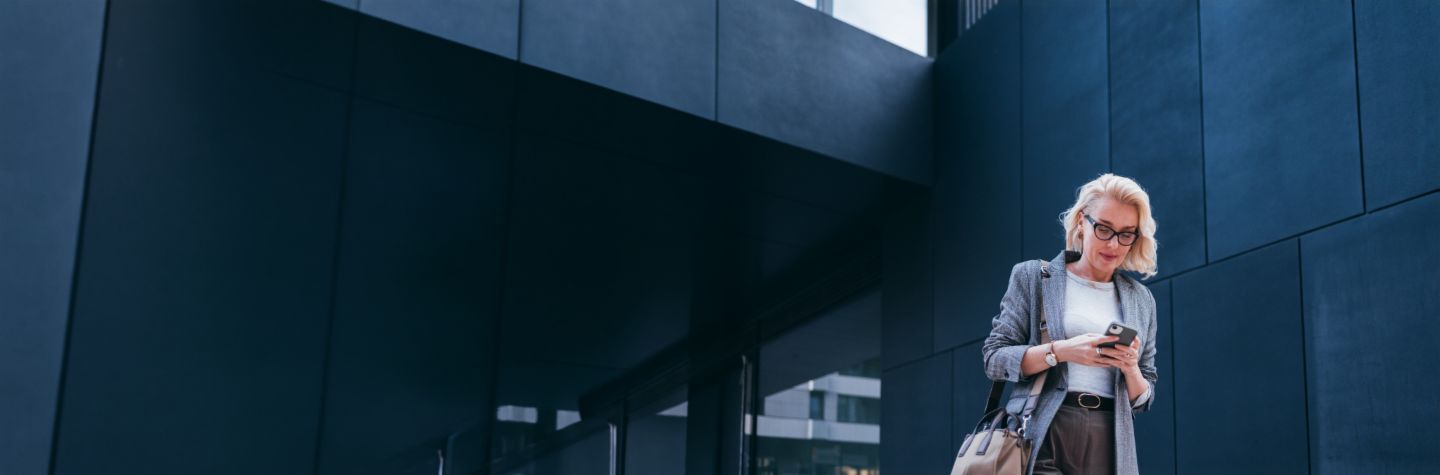 Eine blonde Geschäftsfrau im grauen Blazer steht vor einem modernen Gebäude und schaut lächelnd auf ihr Smartphone.