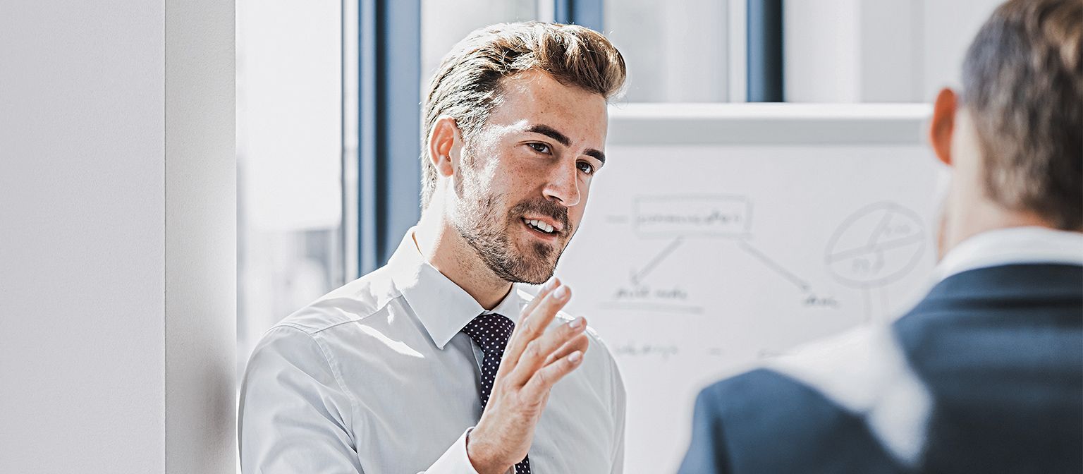 Zu sehen ist ein Mann in Business-Bekleidung, der mit freundlich-engagiertem Ausdruck etwas erklärt. Vor ihm steht eine andere Person, die offensichtlich Zuhörer ist. Im Hintergrund ist ein White-Board mit einer Skizze zu sehen.