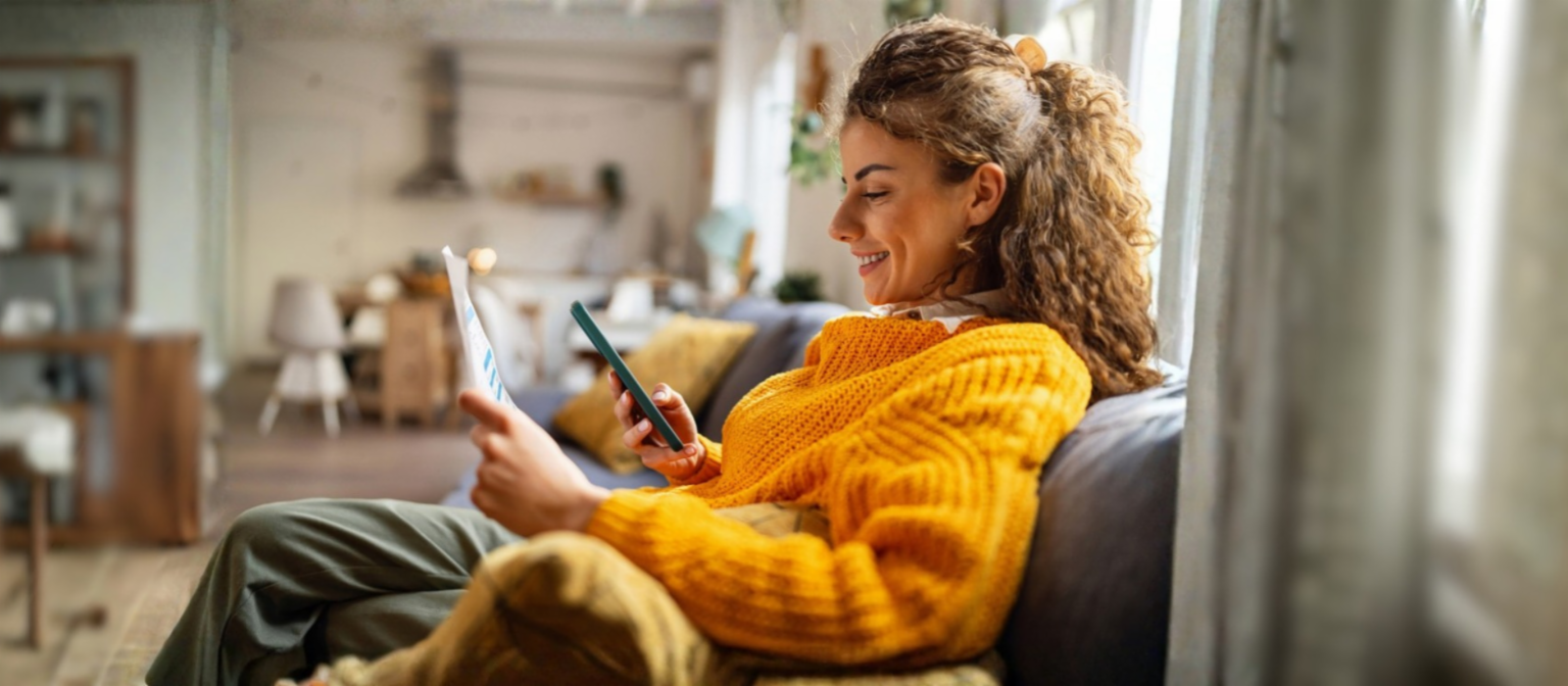 Eine lächelnde junge Frau mit lockigen Haaren in einem gemütlichen gelben Pullover sitzt auf dem Sofa in ihrem Wohnzimmer und schaut auf ihr Smartphone, während sie ein Papierdokument in der Hand hält.
