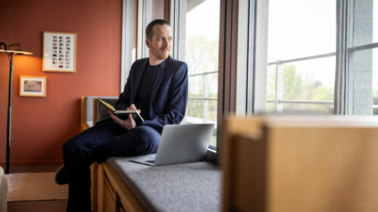 Sebastian Pollmer sitzt mit einem aufgeklappten Laptop und einem Notizbuch in der Hand auf der Fensterbank und schaut aus dem Fenster.