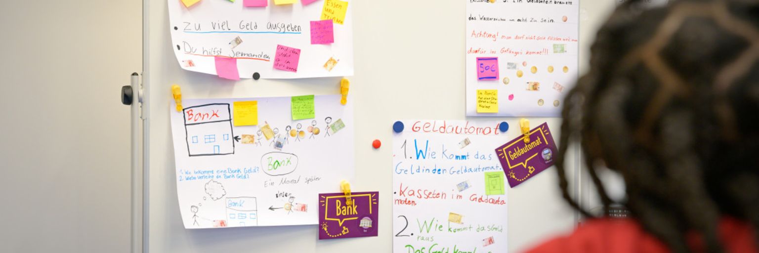 Ein Foto zeigt Post-its und Zettel mit Notizen an einer Tafel, auf denen Begriffe wie „Schulden“, „Bargeld“, „Bank“ und „Geldautomat“ stehen. Darunter steht der Text: „Im Programm ManoMoneta der EOS eigenen finlit foundation können Schüler im Alter von 9 bis 13 Jahren Wissen über Finanzprodukte erwerben.“