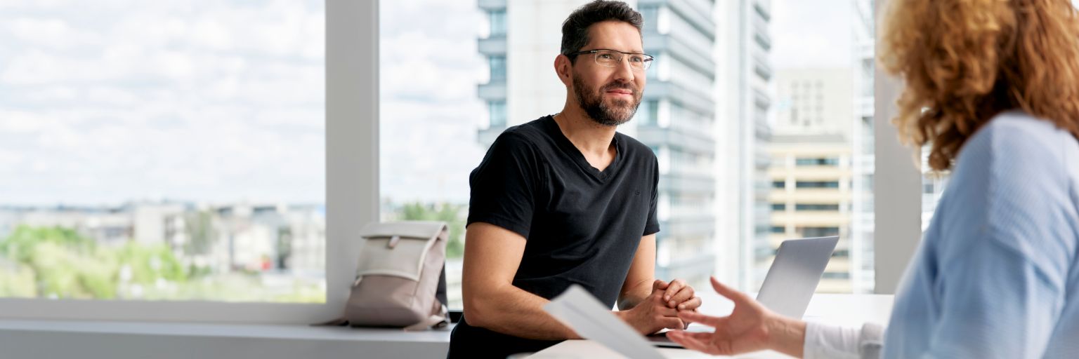 Zwei Personen sitzen in einem modernen, hellen Büroraum und unterhalten sich.<br/>Im Vordergrund sitzt ein lächelnder Mann mit dunklem Haar, Brille und Bart, der ein schwarzes T-Shirt trägt. Vor ihm auf dem Tisch liegen einige Dokumente und ein Laptop. Die zweite Person, von der nur der Oberkörper und die Arme zu sehen sind, sitzt dem Mann gegenüber. Sie trägt eine hellblaue Bluse, hat kurze, krause, rotblonde Haare und hält gestikulierend einige Papiere in der Hand. Im Hintergrund sind große Fenster zu sehen, durch die man auf moderne Gebäude und eine urbane Landschaft blickt.