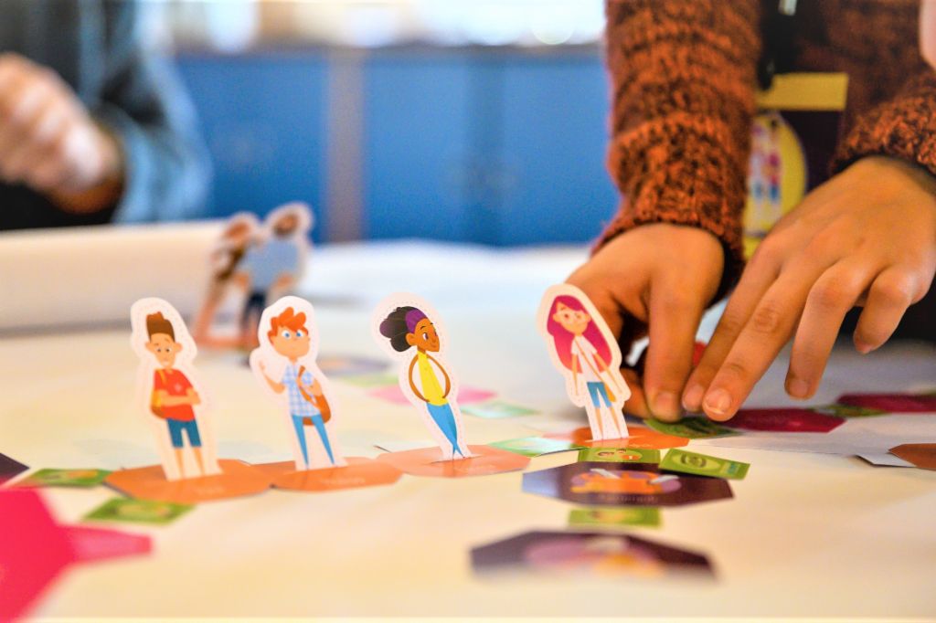 Ein Foto zeigt Hände, die Figuren von Schüler*innen auf einem Spielfeld bewegen. Darunter steht der Text: Finanzbildung: Wer früh den richtigen Umgang mit Geld erlernt, ist später besser vor Überschuldung geschützt.
