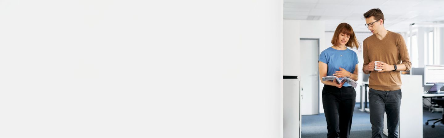 Zwei Kollegen diskutieren in einem modernen Büro, wie EOS im Forderungsmanagement Unternehmen vor Zahlungsausfällen schützen und deren Liquidität maximieren kann, während ein weiterer Mitarbeiter im Hintergrund am Computer arbeitet.