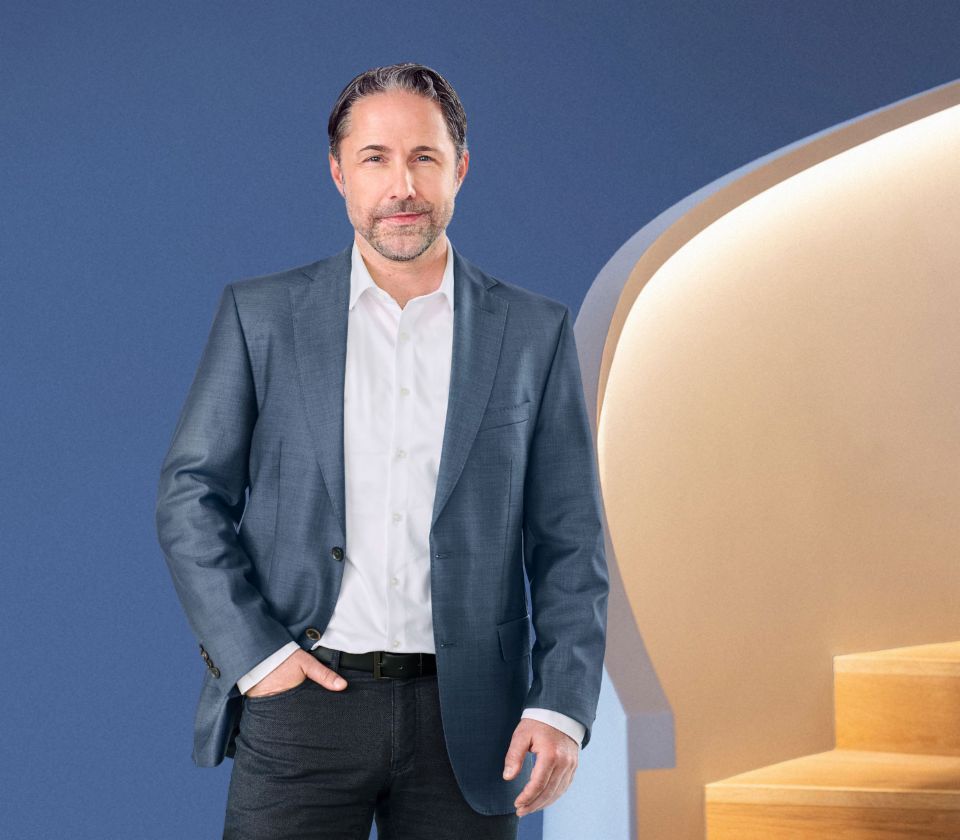 Marwin Ramcke, Vorsitzender der Geschäftsführung der EOS Gruppe steht vor einem blauen Grund und trägt ein dunkles Sakko. 