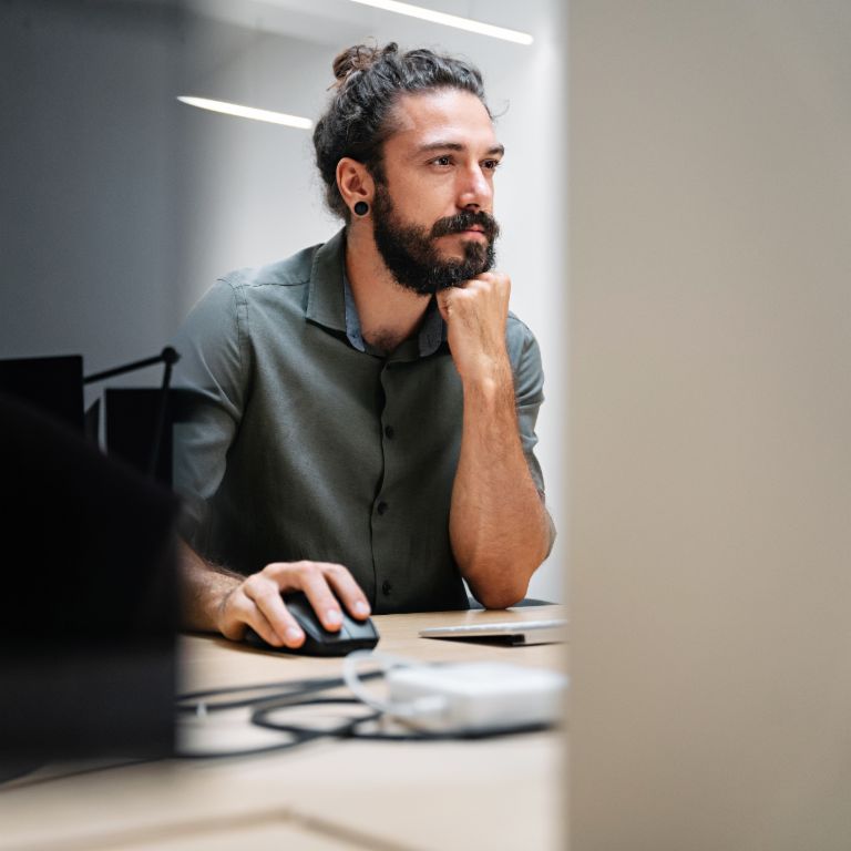 A bearded man with long hair tied in a bun sits at a computer, fully absorbed in his work.