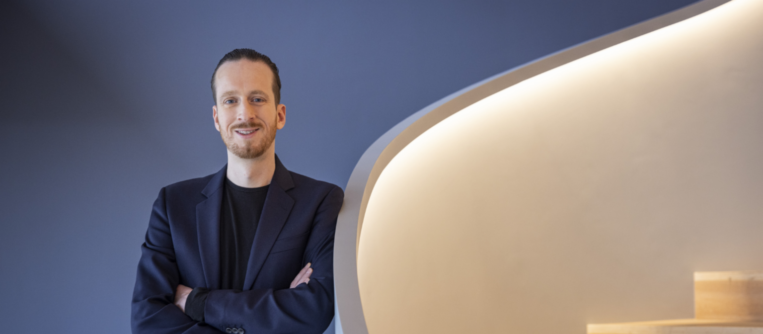 Sebastian Pollmer, ein Mann im blauen Anzug der gegen eine warmweiß beleuchtete Treppe lehnt. 
