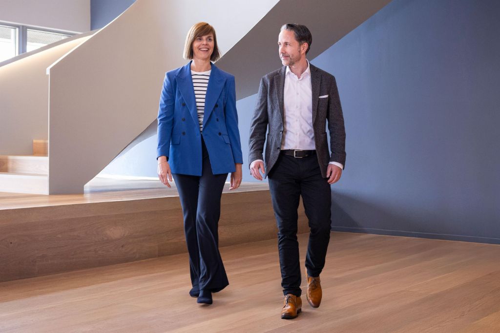 Der CEO im grauen Blazer und die CFO der EOS Gruppe in blauem Blazer gehen in einem modernen Büro. Der Raum hat große Fenster, eine geschwungene Wand und stilvolle hängende Lampen.