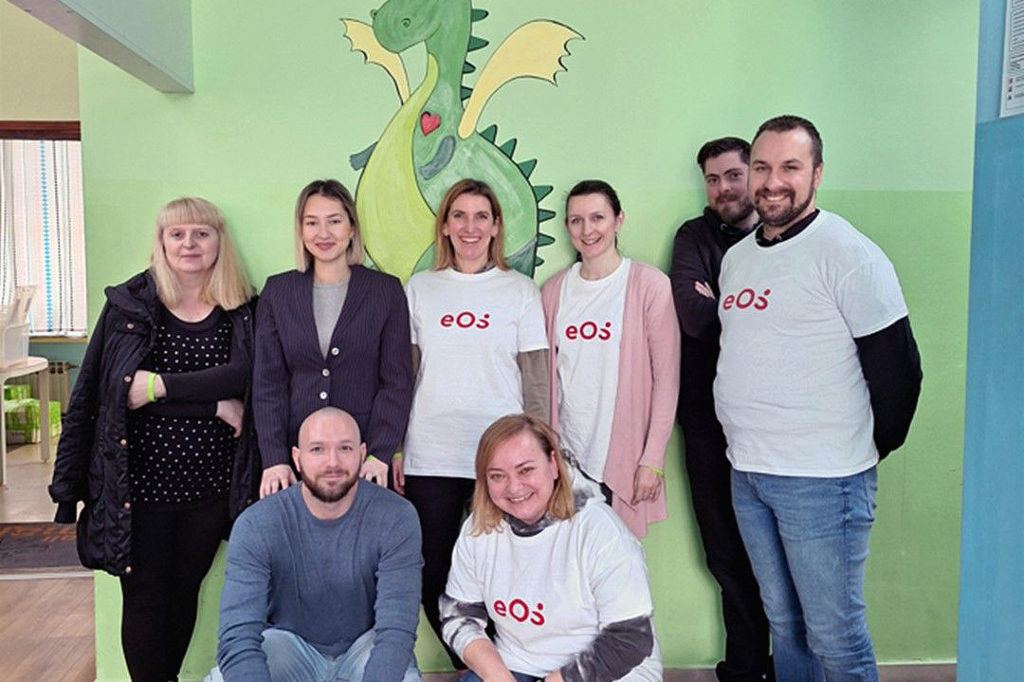 Acht Personen stehen vor einer Wand, auf die ein grüner Drache gemalt ist. Die Personen sind in zwei Reihen angeordnet: Zwei hocken vorne und sechs stehen dahinter. Vier Personen tragen weiße T-Shirts mit dem roten EOS Logo. Die Gruppe lächelt in die Kamera. Auf dem Boden vorne rechts sind einige Flaschen und Getränkekartons zu sehen.