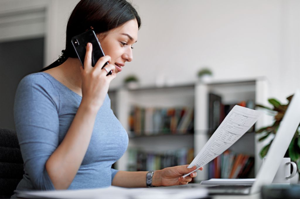 Eine dunkelhaarige, junge Frau in einem blauen Shirt sitzt in einem hellen Raum an einem Tisch mit einem Zettel in der Hand und telefoniert mit ihrem Smartphone vorm Laptop. Im Hintergrund ist ein unscharfes Regal mit Büchern zu sehen.