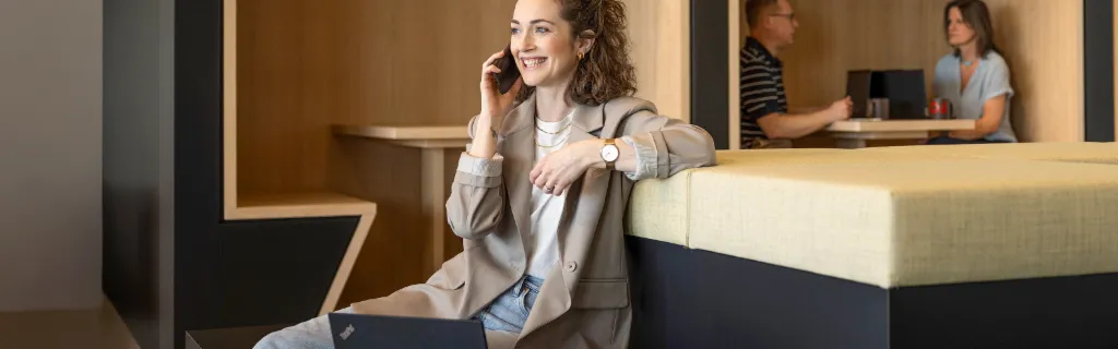 Lächelnde Frau sitzt in einer modernen Büro-Lounge, telefoniert und arbeitet am Laptop. Im Hintergrund arbeiten zwei weitere Personen.