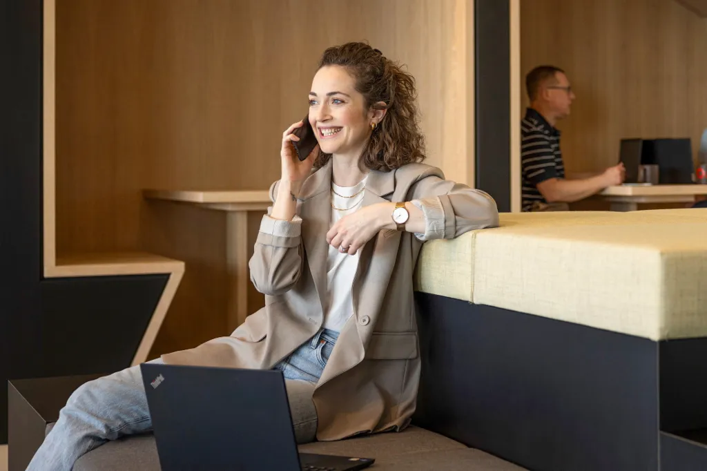 Lächelnde Frau sitzt in einer modernen Büro-Lounge, telefoniert und arbeitet am Laptop. Im Hintergrund arbeiten zwei weitere Personen.